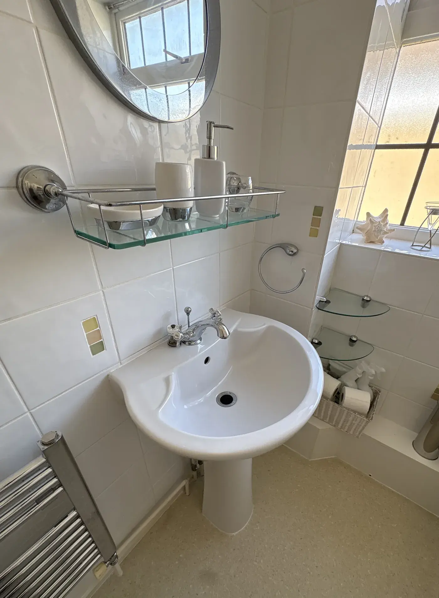 White tiled bathroom with pedestal basin, glass shelves and chrome towel rail, all wiped down