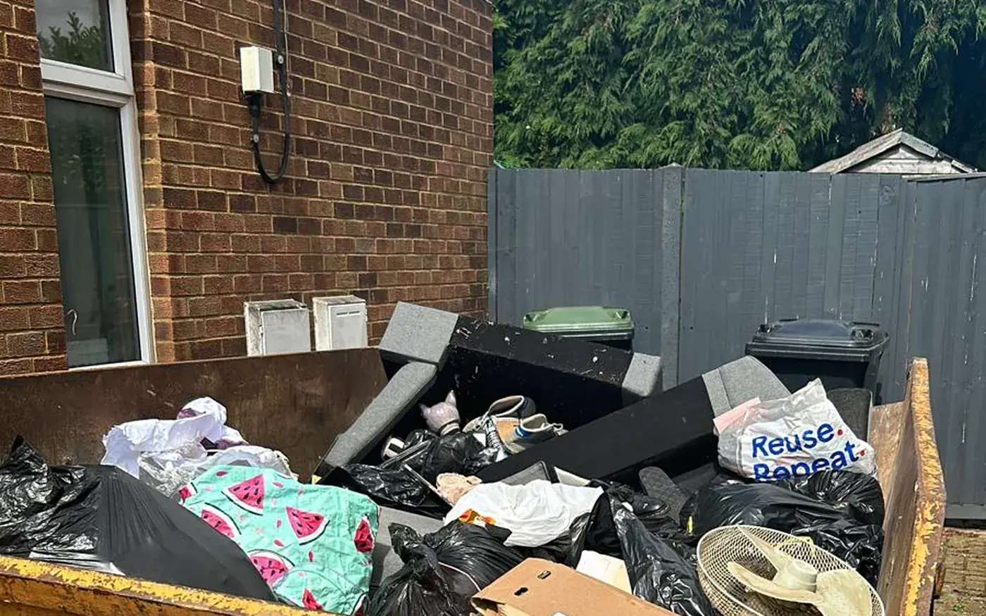A skip beside a house filled with bagged waste, a mattress and old furniture