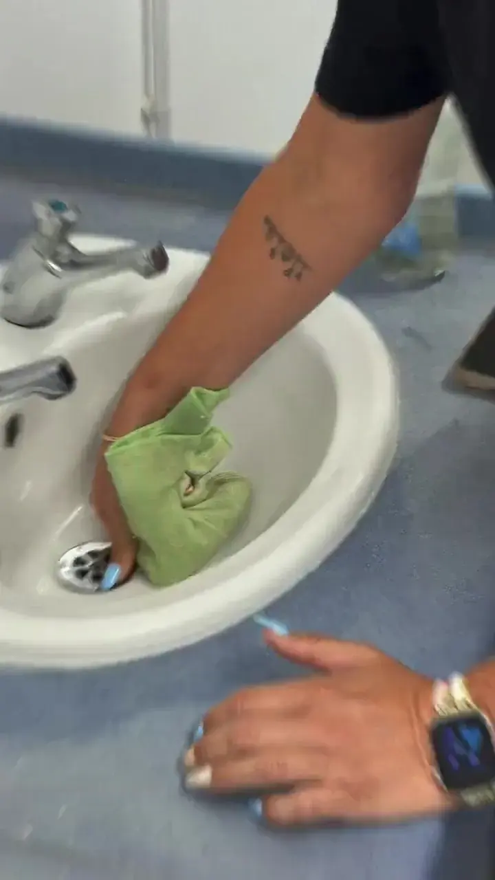 Hands wiping around a white washroom basin and plughole with a green cloth