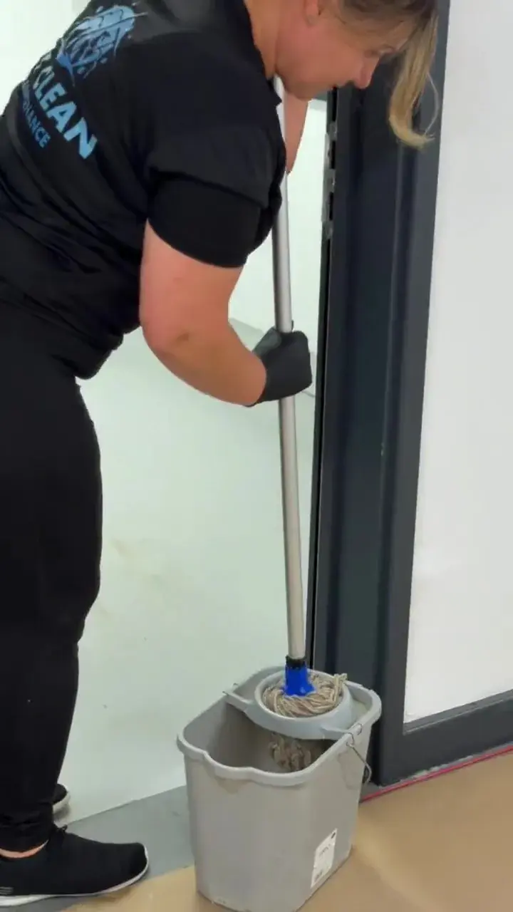 A cleaner in branded uniform wringing a mop into a bucket on a hard floor