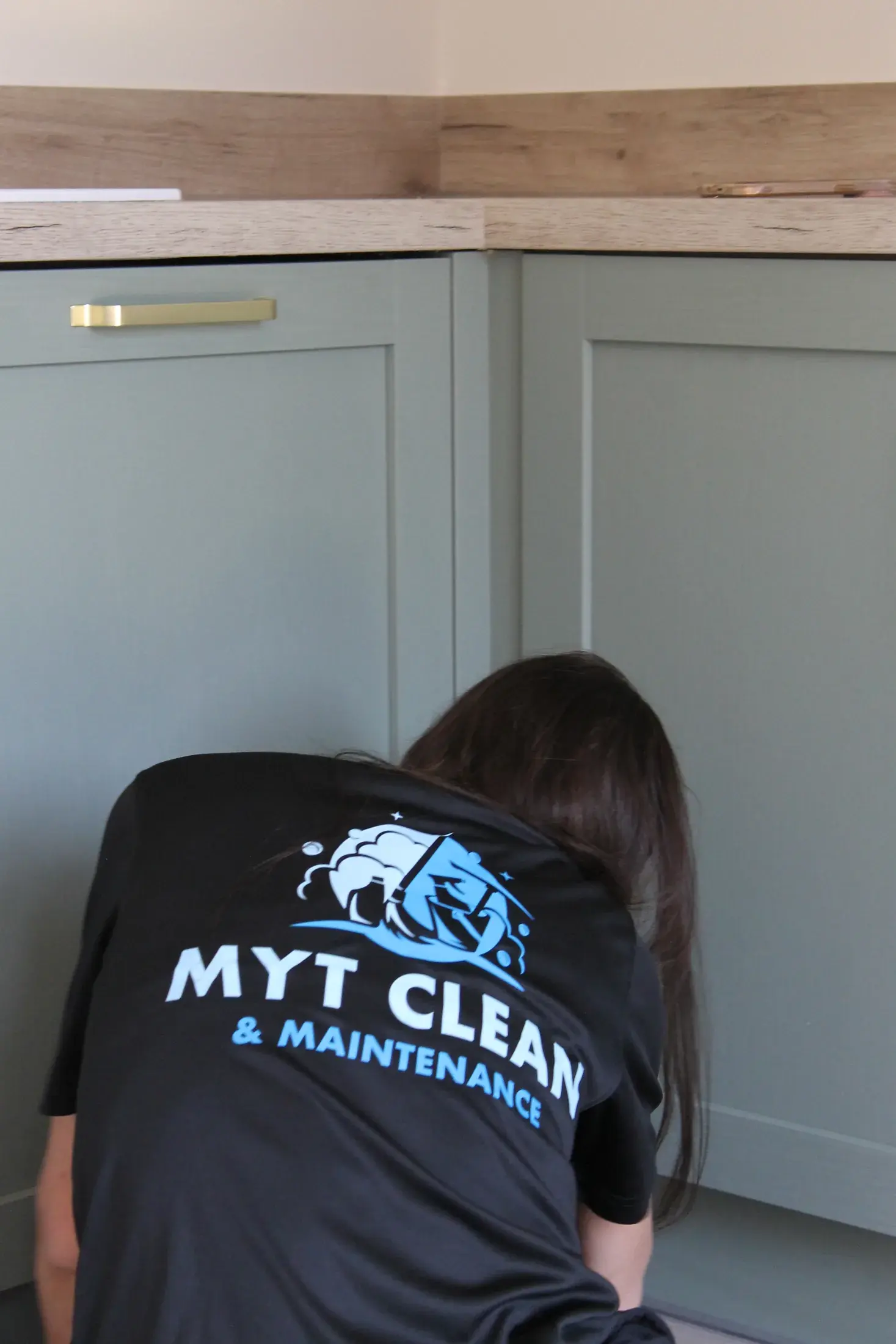 A worker in a MyT Clean and Maintenance shirt cleaning sage green kitchen units